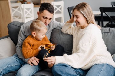 Mutlu beyaz ebeveynler ve küçük oğlu evde kanepede otururken gülümseyip oyuncaklarla oynuyorlar.