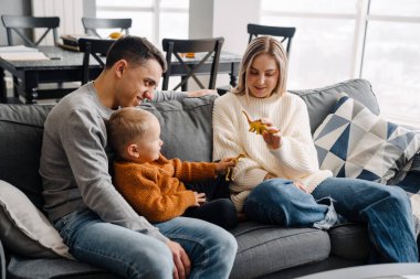 Mutlu beyaz ebeveynler ve küçük oğlu evde kanepede otururken gülümseyip oyuncaklarla oynuyorlar.