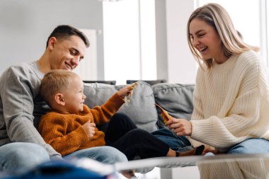 Mutlu beyaz ebeveynler ve küçük oğlu evde kanepede otururken gülümseyip oyuncaklarla oynuyorlar.