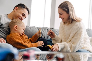 Mutlu beyaz ebeveynler ve küçük oğlu evde kanepede otururken gülümseyip oyuncaklarla oynuyorlar.