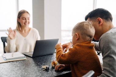 Genç beyaz kadın dizüstü bilgisayar kullanırken adam ve küçük oğlu evde oyuncaklarla oynuyordu.