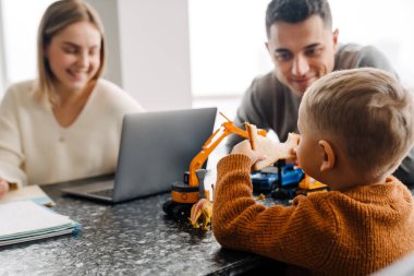 Genç beyaz kadın dizüstü bilgisayar kullanırken adam ve küçük oğlu evde oyuncaklarla oynuyordu.