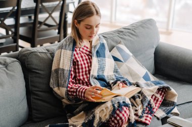 Evdeki kanepede otururken battaniyeye sarılı kitap okuyan mutlu genç bir kadın.