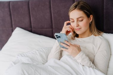 Evde yatarken gülümseyen ve cep telefonu kullanan genç beyaz sarışın kadın.