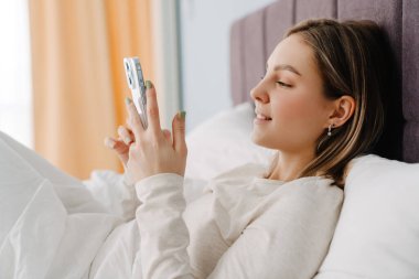 Evde yatarken gülümseyen ve cep telefonu kullanan genç beyaz sarışın kadın.