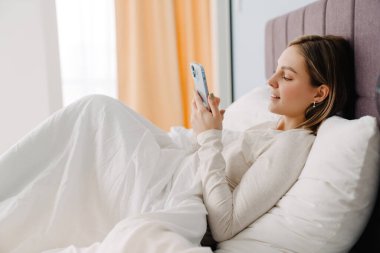 Evde yatarken gülümseyen ve cep telefonu kullanan genç beyaz sarışın kadın.