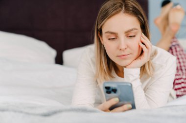 Genç beyaz sarışın kadın evde yatarken cep telefonu kullanıyor.