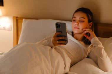 Genç beyaz sarışın kadın evde yatarken cep telefonu kullanıyor.