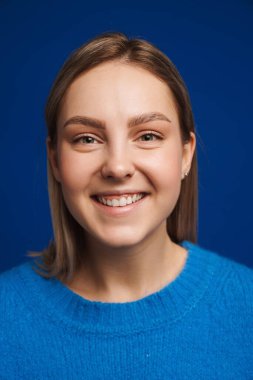 Genç, güzel, mutlu, gülümseyen bir kızın portresi mavi izole arka planda kameraya bakıyor.