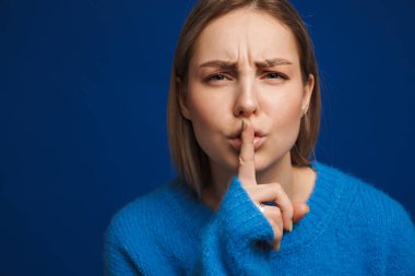 Süveter giyen, kaşlarını çatan ve mavi arka planda izole edilmiş sessiz hareketler yapan genç beyaz kadın.