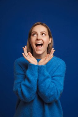 Genç, mutlu, süveter giyen, mavi arka planda tek başına duran genç bir kadın.