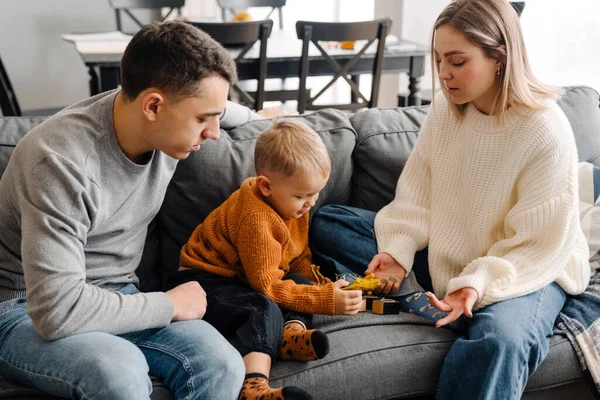 Mutlu beyaz ebeveynler ve küçük oğlu evde kanepede otururken gülümseyip oyuncaklarla oynuyorlar.