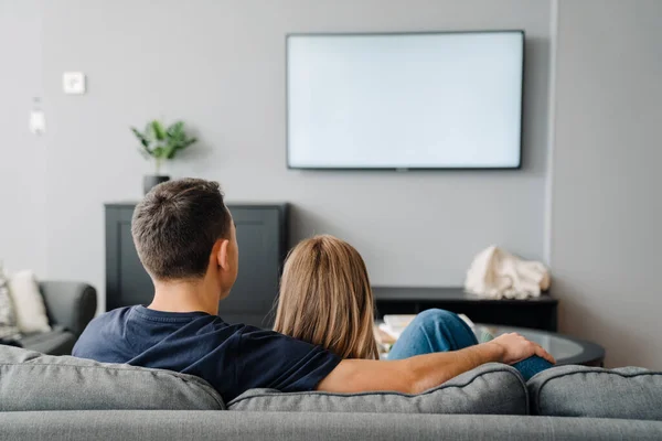 Young white man and woman hugging while sitting on sofa and watching TV at home