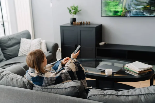 Evdeki kanepede televizyon izlerken battaniyeye sarılı genç, mutlu bir kadın.