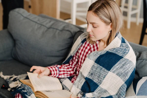 Evdeki kanepede otururken battaniyeye sarılı kitap okuyan mutlu genç bir kadın.