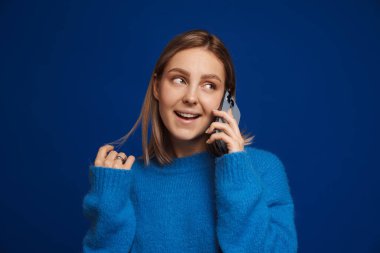 Genç ve çekici bir kız, mavi izole edilmiş arka planda telefonla konuşurken açık ağzıyla saçlarıyla oynuyor.