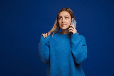 Genç ve çekici kız saçlarıyla oynuyor, mavi izole edilmiş arka planda telefonla konuşurken dudağını ısırıyor.