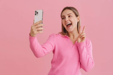 Genç güzel beyaz kadın gülümsüyor ve cep telefonuyla selfie çekiyor. Pembe arka planda tek başına duruyor.