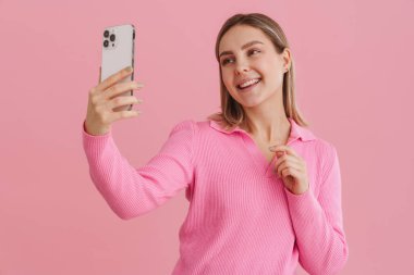 Genç, güzel, gülümseyen mutlu kız pembe kazaklı selfie çekiyor izole edilmiş mavi arka planda