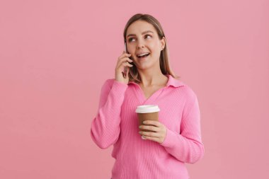 Genç, güzel, beyaz bir kadın kahve fincanı tutuyor ve cep telefonuyla konuşuyor. Pembe arka planda tek başına duruyor.