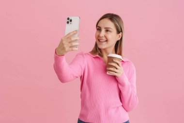 Genç, güzel, beyaz bir kadın kahve fincanı tutuyor ve cep telefonuyla selfie çekiyor. Pembe arka planda tek başına duruyor.
