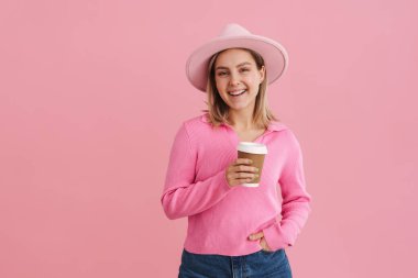 Pembe şapkalı ve kazaklı genç bir bayan kameraya bakıyor ve pembe arka planda duruyor.