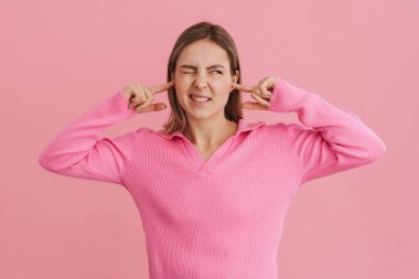 Genç, güzel, beyaz bir kadın, kaşlarını çatmış, kulaklarını tıkamış pembe arka planda tek başına dikiliyor.