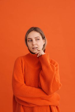 Turuncu kazaklı genç bayan çenesine dokunuyor ve izole edilmiş turuncu arka planda sağına bakıyor.