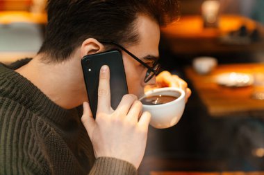Genç beyaz adam gözlük takıyor kahve içiyor ve kafede cep telefonu kullanıyor.
