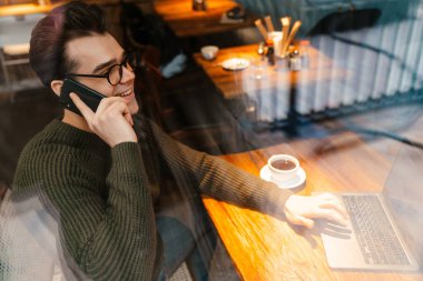 Kahve içerken ve kafede cep telefonu kullanırken gülümseyen gözlüklü genç bir beyaz adam.