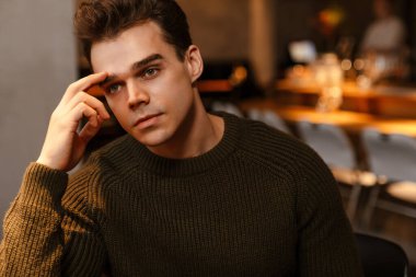 Süveter giyen genç, beyaz, esmer bir adam kafeye bakıyor ve düşünüyor.