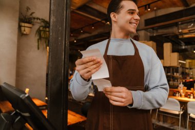 Young white waiter man wearing apron standing in front of computer and holding client receipt in cafe clipart