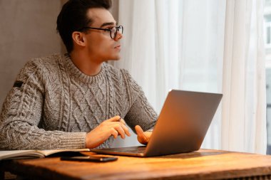 Gözlüklü genç beyaz adam laptopta çalışıyor ve kafede otururken notlar alıyor.