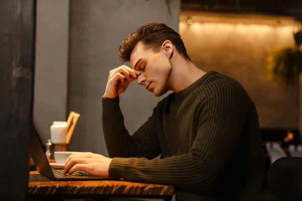 在咖啡店使用笔记本电脑时 年轻的白人穿着毛衣 看起来很疲倦 — 图库照片