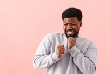 Siyah, sakallı bir adam pembe arka planda yalnız poz verirken yüzünü buruşturuyor.