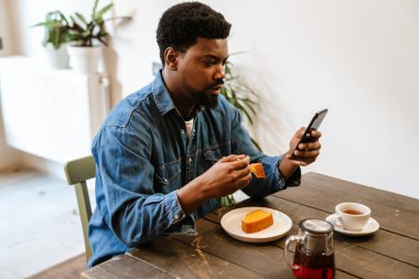 Kafede kek yerken cep telefonu kullanan siyah sakallı bir adam.