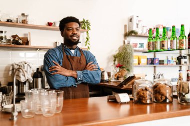 Kafede çalışırken önlük takan siyah sakallı adam gülümsüyor.