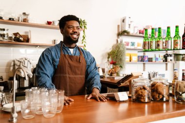 Kafede çalışırken önlük takan siyah sakallı adam gülümsüyor.