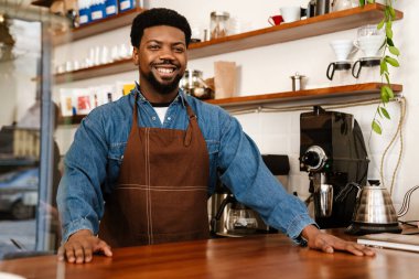 Kafede çalışırken önlük takan siyah sakallı adam gülümsüyor.