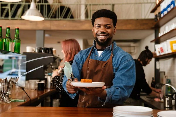Black bearded man wearing apron smiling while working in cafe indoors