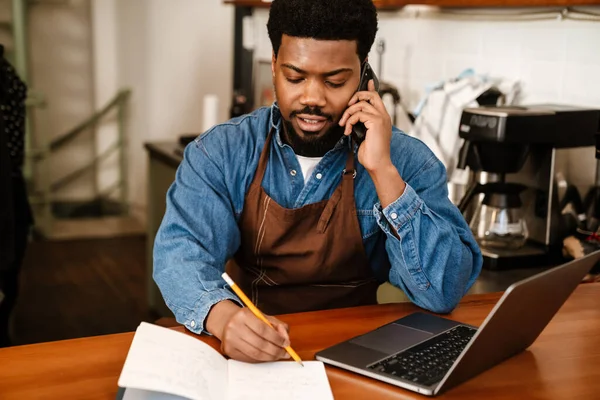 Black bearded man wearing apron talking on cellphone while working in cafe indoors