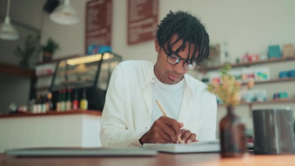 カフェでノートに何かを書いている恐ろしいロックを持つ幸せなアフリカ人男性 — ストック動画