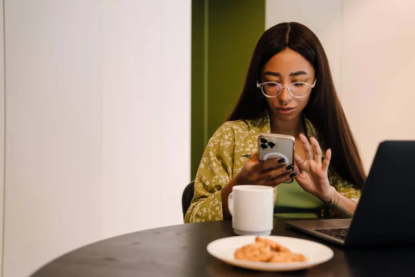 Młoda Piękna Kobieta Okularach Patrząc Telefon Podczas Pracy Laptopie Filiżanką — Zdjęcie stockowe