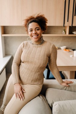 Genç, güzel, gülümseyen Afrikalı kadın evdeki kanepede oturan kameraya bakıyor.
