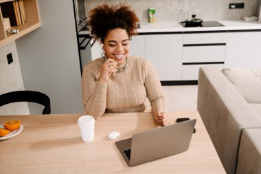 Genç, mutlu, gülümseyen, elinde kahve ve dizüstü bilgisayarıyla evde video konferansı düzenleyen Afrikalı bir kadın.