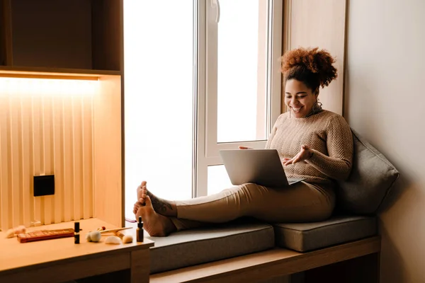 Young beautiful happy smiling african woman with laptop holding video conference sitting on windowsill