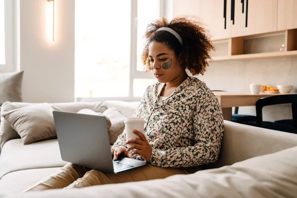 若いアフリカ系アメリカ人の女性はコーヒーを飲みながら家でソファに座っている間にノートパソコンを使う — ストック写真