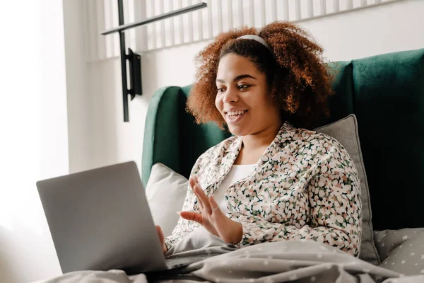 若いアフリカ系アメリカ人の女性は笑顔と自宅でベッドに横たわっている間 ラップトップを使用して — ストック写真