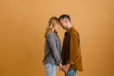 Bej arka planda el ele tutuşurken gülümseyen beyaz mutlu çift.