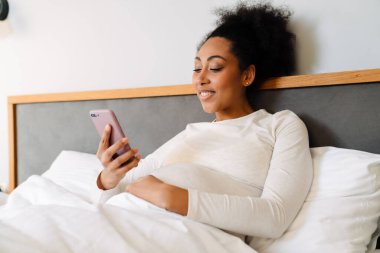 Young smiling beautiful pregnant afro woman liying on bed and looking on her phone in cozy bedroom at home